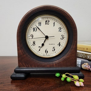 May include: A wooden quartz clock with a cream-coloured face and black numerals. The clock has a dark brown arched frame with a black base. The clock face reads "QUARTZ" and "MADE IN FRANCE".