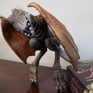 May include: A detailed figurine of a dragon with a menacing open mouth, sharp teeth, and large, outstretched wings. The dragon is primarily gray and brown, with orange wings. The figurine is posed on a wooden surface, with a book visible in the background.