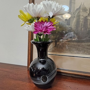 May include: A black glass vase with a white swirl design holds a bouquet of white, yellow, and magenta chrysanthemums. The vase sits on a wooden surface, with a framed picture of a city in the background.