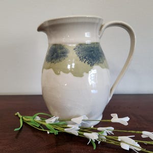 Puede incluir: Jarra de cerámica blanca con asa y boquilla. La jarra tiene una banda de diseños florales verdes y azules en el centro. Una ramita de flores blancas descansa frente a la jarra.