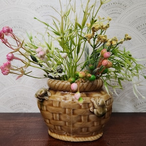 Puede incluir: Un jarrón de cerámica con forma de cesta y textura tejida, adornado con dos figuras de rana. El jarrón contiene un arreglo de flores artificiales, incluyendo flores rosas, verdes y amarillas, creando una exhibición decorativa.