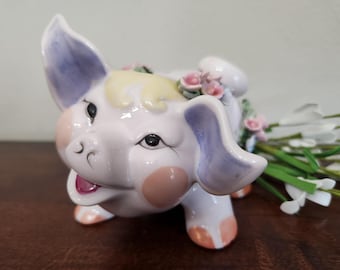 May include: A ceramic pig ornament with a pale pink body, lilac ears, and peach accents. The pig has a smiling face and is decorated with a floral crown and small pink flowers. White flowers and green stems are in the background.