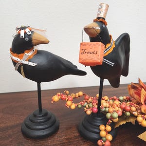May include: Two black crow ornaments with orange and white detailing, ideal for Halloween. One crow wears a veil, the other a top hat and carries a small orange bag that says "Treats". They stand on black bases.