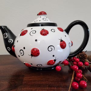 Puede incluir: Una tetera de cerámica blanca decorada con mariquitas rojas, espirales negras y puntos negros. El pico y el asa son negros. La tapa tiene una mariquita encima. Un artículo decorativo para la cocina.