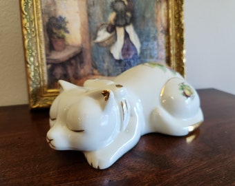 May include: A white ceramic cat figurine in a sleeping pose, with gold accents on the ears and paws. The cat has closed eyes and a serene expression. Decorative fruit and leaves are painted on the side. The figurine is on a dark wooden surface.
