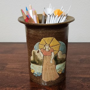 以下が含まれることがあります： 傘を持った女性の装飾的なイメージが描かれた、茶色のセラミック製のペン立て。ペン立てには色鉛筆と筆が入っています。ペン立ては木製の表面に置かれています。