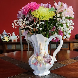 May include: A white ceramic pitcher with a seashell design holds a vibrant bouquet of flowers. The flowers include pink, purple, and yellow blooms, with white baby's breath. The pitcher sits on a dark wooden table, creating a colorful centerpiece.