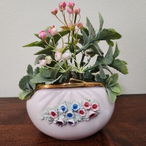 Può includere: Un piccolo vaso in ceramica rosa chiaro con bordo dorato, riempito di fiori e verde artificiali. Il vaso presenta un rilievo floreale decorativo nei toni del blu, rosso e viola. La composizione comprende boccioli rosa e foglie verdi.