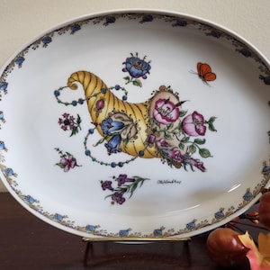 Puede incluir: Plato ovalado para servir con fondo blanco y borde decorativo. El plato presenta un diseño de cuerno de la abundancia lleno de flores, una mariposa y detalles de cuentas. El cuerno de la abundancia es amarillo, azul y rosa.