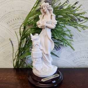 Puede incluir: Figura de porcelana blanca de una mujer sosteniendo dos palomas. La figura está sobre una base de madera oscura con una etiqueta dorada que dice "Giuseppe Armani". La mujer lleva un vestido fluido. La figura mide aproximadamente 25 cm de altura.