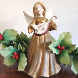 Può includere: Statuina di angelo in ceramica con abito dorato e ali bianche, che suona un mandolino. L'angelo è circondato da una ghirlanda di foglie verdi e bacche rosse, creando una scena festosa natalizia.