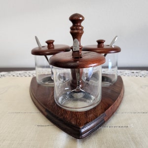 May include: A vintage condiment set featuring three clear glass jars with wooden lids and small spoons. The jars are set on a dark wooden triangular base with a central handle. The set is designed for serving condiments.