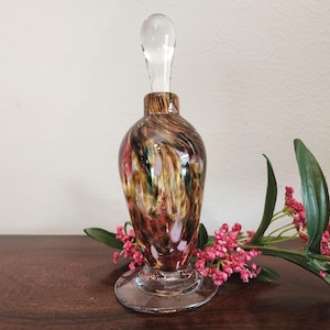 May include: A decorative glass perfume bottle with a clear stopper. The bottle has a bulbous shape with a clear base and is decorated with swirls of brown, gold, red, and white. Pink flowers and green leaves are next to the bottle.