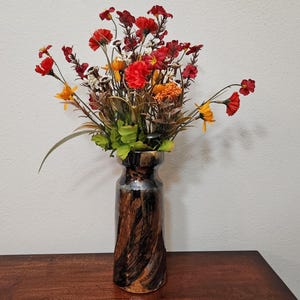Könnte beinhalten: Eine dekorative Vase mit einem braunen und schwarzen Wirbelmuster enthält ein Arrangement künstlicher Blumen. Die Blumen sind rot, gelb und orange, mit grünen Blättern. Die Vase steht auf einer Holzoberfläche.