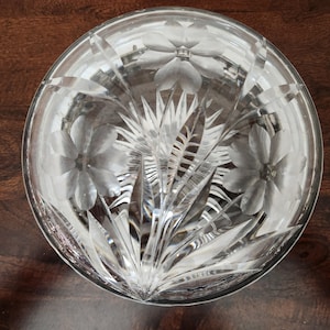 May include: A clear glass bowl with etched floral and leaf designs. The bowl features three large flowers and a central arrangement of leaves. The glass is transparent, allowing light to pass through, and is likely used for serving or display.