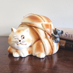 May include: A vintage ceramic cat-shaped cookie jar with a lid, featuring a playful design. The cat is white and orange, with a smiling face. A small gray mouse is perched on the side. The jar is a decorative kitchen item.