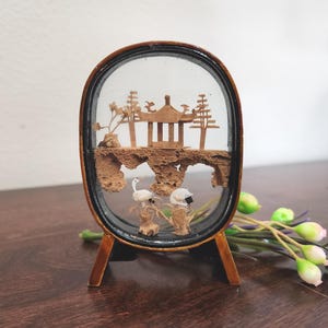 May include: An oval-shaped decorative piece featuring a miniature Asian-inspired landscape. The diorama includes a wooden pagoda, trees, and two white birds, all encased in glass and set on a wooden stand. The frame is dark brown.