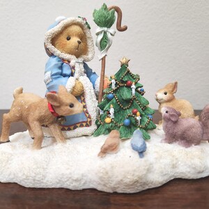 May include: A Christmas figurine featuring a teddy bear in a blue coat, a decorated Christmas tree, a deer with a red collar, and several small animals on a snowy base. The figurine includes a staff with a green top.
