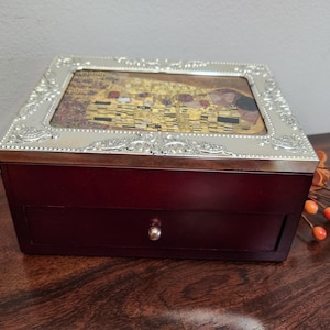 May include: A rectangular, dark red wooden jewelry box with a silver-colored, ornate frame. The top features a print of Gustav Klimt's "The Kiss." The box has a small drawer with a silver knob.