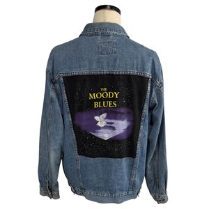 May include: A light blue denim jacket. The back features a black patch with the text "THE MOODY BLUES" and a white bird graphic. The jacket has a classic design with button closures and pockets.