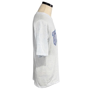 May include: Light gray t-shirt with a blue and white graphic on the sleeve. The shirt is displayed on a mannequin. The graphic appears to be a stylized letter.