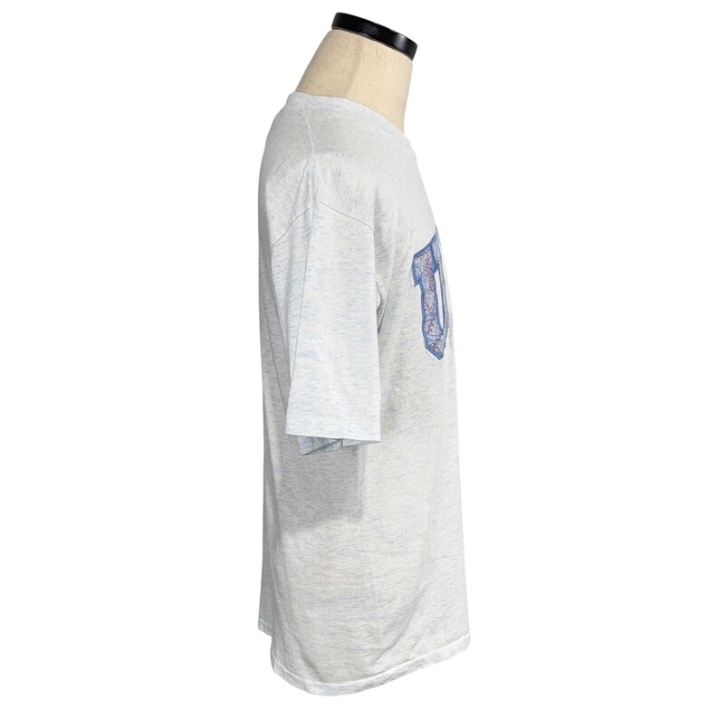 May include: Light gray t-shirt with a blue and white graphic on the sleeve. The shirt is displayed on a mannequin. The graphic appears to be a stylized letter.