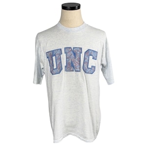May include: Light gray t-shirt with the letters "UNC" in blue with a paisley design. The shirt is displayed on a mannequin. The t-shirt has short sleeves and a crew neck.