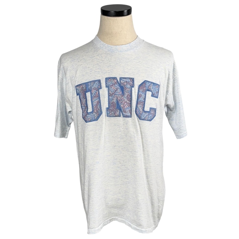 May include: Light gray t-shirt with the letters "UNC" in blue with a paisley design. The shirt is displayed on a mannequin. The t-shirt has short sleeves and a crew neck.
