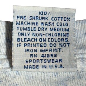 May include: A close-up of a white fabric tag with blue text. The tag reads "100% PRE-SHRUNK COTTON MACHINE WASH COLD. TUMBLE DRY MEDIUM. ONLY NON-CHLORINE BLEACH ON COLORS. IF PRINTED DO NOT IRON IMPRINT. RN 41253 SPORTSWEAR MADE IN U.S.A."