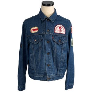 May include: A vintage blue denim jacket with multiple patches. Patches include "TWA" and "Technical Excellence Continental Airlines 1986". The jacket has a button-up front, two chest pockets, and a classic collar. A red Levi's tag is visible.