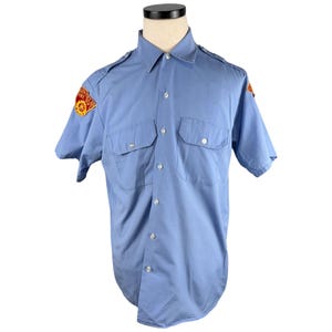Può includere: Camicia azzurra a maniche corte con due tasche sul petto e chiusura con bottoni. Presenta spalline e toppe con testo e un emblema rosso e giallo. La camicia è esposta su un manichino.
