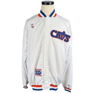 May include: White satin jacket with blue and orange trim. Features the word "CAVS" in blue and orange lettering on the front. Includes a small NBA logo and a "Basketball Centennial" patch. The jacket has a snap-front closure.
