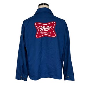 Puede incluir: Chaqueta azul con un parche con el logo de Miller High Life rojo y blanco en la espalda. La chaqueta tiene una botonadura frontal, mangas largas y un cuello. La chaqueta está hecha de un material duradero.