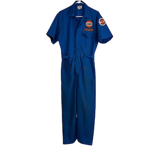 May include: A blue short-sleeve jumpsuit with a collared neckline and a front zipper closure. The jumpsuit features two orange and white logo patches, one on the chest and one on the sleeve. The text "CLARK" and "The Performers" are visible on the patches.