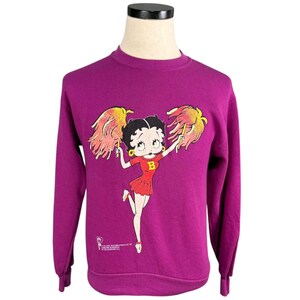 May include: A magenta sweatshirt featuring a Betty Boop graphic. The cartoon character is depicted in a red cheerleader outfit with yellow pom-poms. The sweatshirt has long sleeves and a crew neck. The graphic is centered on the front.