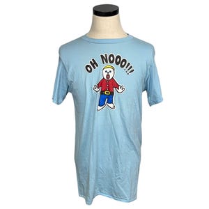 May include: Light blue t-shirt featuring a cartoon character with the text "OH NOOO!!!". The character has a surprised expression, red shirt, blue pants, and white shoes. The shirt is displayed on a mannequin.
