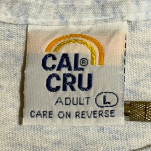 May include: Close-up of a white fabric tag with the text "CAL CRU" in dark blue, and "ADULT L" in black. The tag has a gold and orange arched design at the top and the words "CARE ON REVERSE" at the bottom.