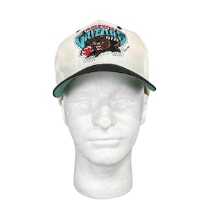 Puede incluir: Gorra de béisbol blanca con visera negra y un logotipo azul y verde de los Vancouver Grizzlies con un oso. El logotipo dice "Vancouver Grizzlies" y el oso tiene un balón de baloncesto en su pata.