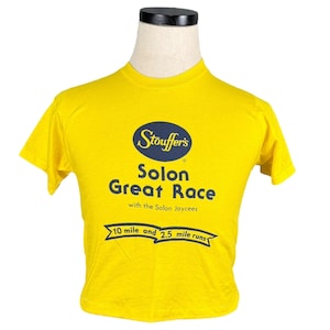 May include: A yellow t-shirt with the text "Stouffer's Solon Great Race" in navy blue. The shirt also features text indicating 10-mile and 2.5-mile runs. The shirt is displayed on a mannequin.
