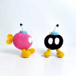 May include: Two crocheted Bob-omb characters, one pink and one black, with white eyes, yellow feet, and a blue and white fuse. The pink Bob-omb has yellow wings. The characters are handmade and sit on a white surface.