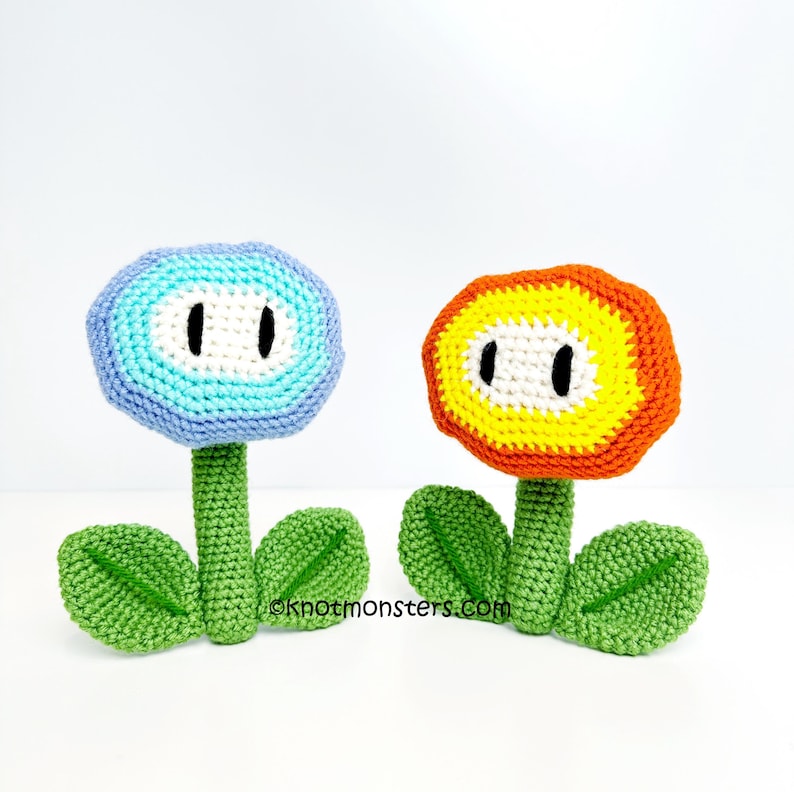 May include: Two crocheted flower figures. One is blue with a white center and black eye details, the other is orange and yellow with a white center and black eye details. Both have green stems and leaves. The website address oknotmonsters.com is visible.