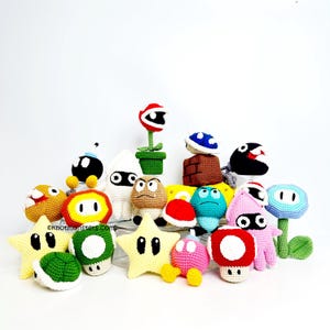 Può includere: Una collezione di personaggi di videogiochi colorati all'uncinetto, tra cui stelle, funghi e una pianta Piranha. I personaggi sono in vari colori, come giallo, rosso, verde e blu, e sono disposti su uno sfondo bianco. L'indirizzo del sito web "oknotmonsters.com" è visibile.