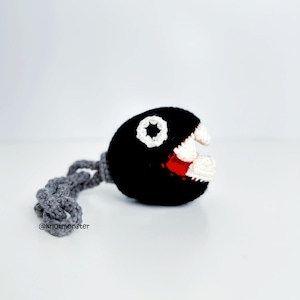 May include: A crocheted Chain Chomp character from the Super Mario video game series. The black ball has a white star, white teeth, and a red mouth. A gray chain extends from the ball. The text "@knotmonster" is visible.