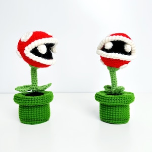 May include: Two crocheted Piranha Plants from the Super Mario video game series. Each plant features a red head with white spots and a white-lined mouth, black interior, green stem, and green pot. The base of each pot has the text "knotmonsters.com".