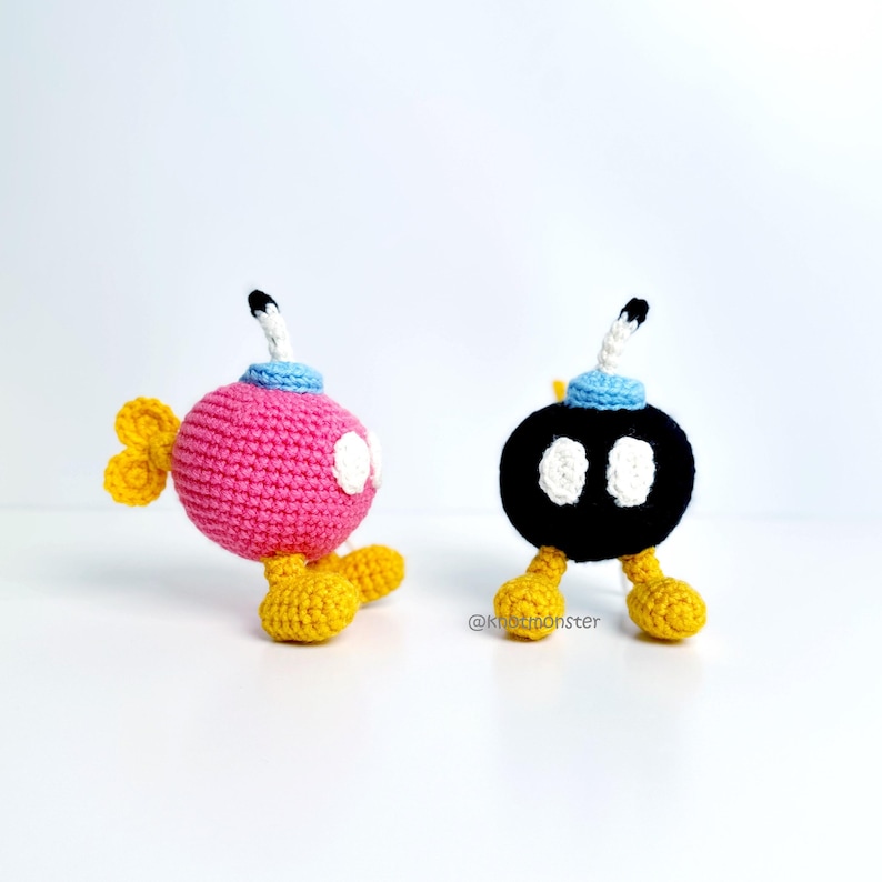 May include: Two crocheted Bob-omb characters, one pink and one black, with white eyes, yellow feet, and a blue and white fuse. The pink Bob-omb has yellow wings. The characters are handmade and sit on a white surface.