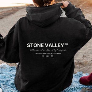 May include: Black Stone Valley hoodie with white text on the back. The text reads "STONE VALLEY" and includes the phrases "SLOW DOWN, RELAX & ENJOY LIFE A LITTLE MORE." The person is holding a slice of pizza.