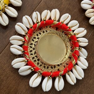 May include: A round, gold-coloured metal tray with a red and gold trim. The tray is decorated with white seashells around the edge.
