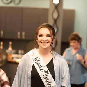 Puede incluir: Una mujer con una bata azul claro con una banda blanca que dice "Brooke Bridesmaid" en letras negras. Ella est&aacute; sonriendo y mirando a la c&aacute;mara.