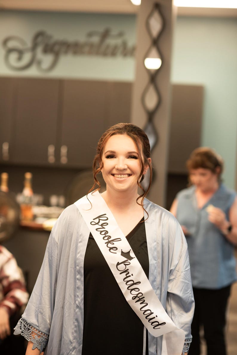 Puede incluir: Una mujer con una bata azul claro con una banda blanca que dice "Brooke Bridesmaid" en letras negras. Ella est&aacute; sonriendo y mirando a la c&aacute;mara.