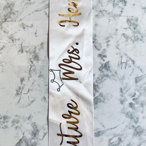 Puede incluir: Banda de sat&eacute;n blanco con letras doradas que dicen "Future Mrs. [Last Name]" con una corona y un gr&aacute;fico de anillo de diamantes.